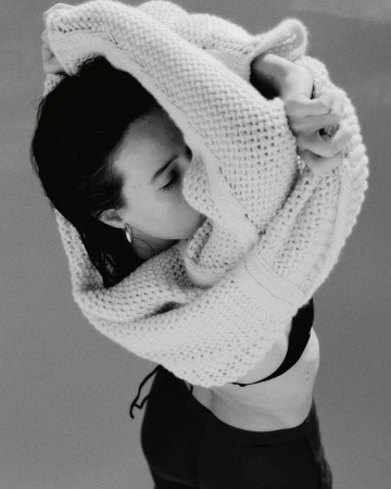 Monochrome portrait of a woman in a black top and cardigan, gazing down. The shot is from above with soft lighting and minimal background.