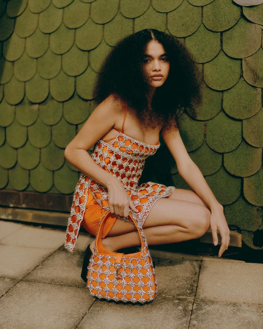 A woman in an orange dress poses with a handbag outdoors against a textured green wall on a rooftop, creating a fashion-editorial aesthetic.