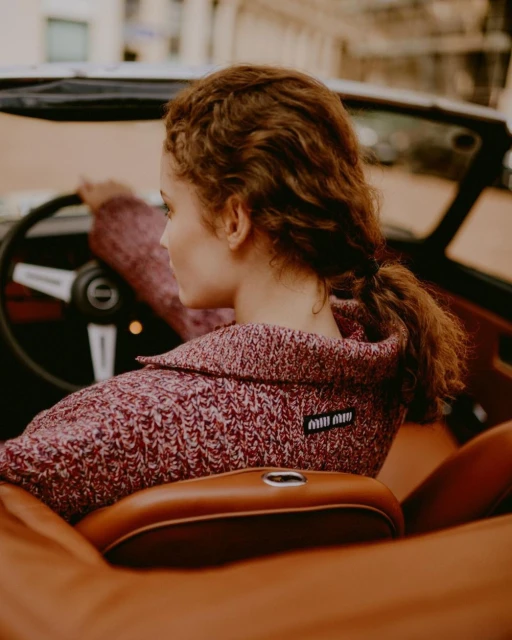 Woman driving a vintage convertible car with a reddish-brown knit sweater, seated in tan leather interior. The Miu Miu logo is visible on her sweater.
