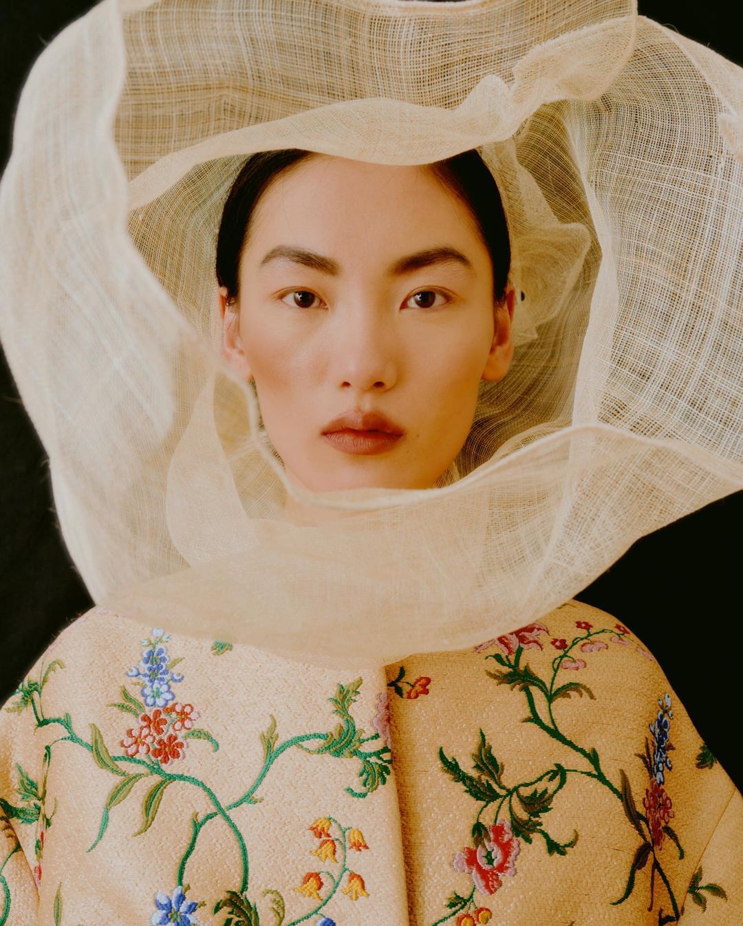 Portrait of Asian woman with a neutral facial expression wearing a sheer fabric headpiece and floral-patterned embroidered jacket.