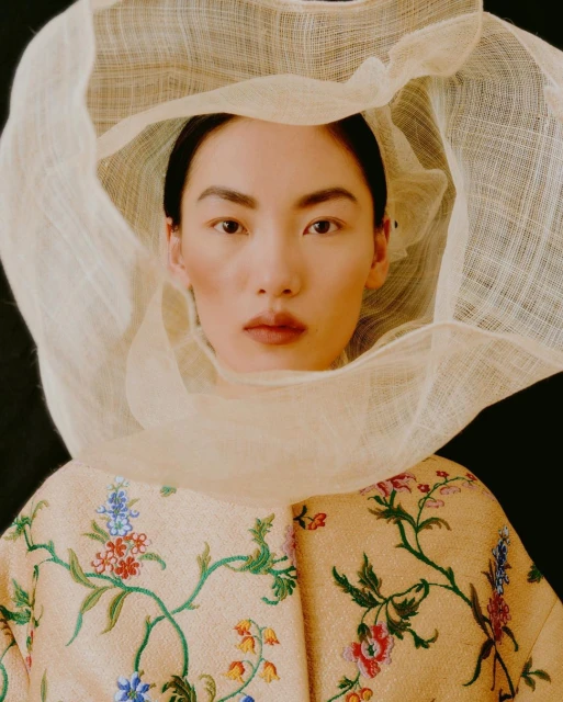 Portrait of Asian woman with a neutral facial expression wearing a sheer fabric headpiece and floral-patterned embroidered jacket.
