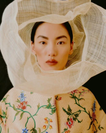 Portrait of Asian woman with a neutral facial expression wearing a sheer fabric headpiece and floral-patterned embroidered jacket.
