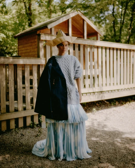 Woman poses in nature wearing a paper hat, cable-knit sweater, layered blue dress and jacket against a wooden fence and shed.