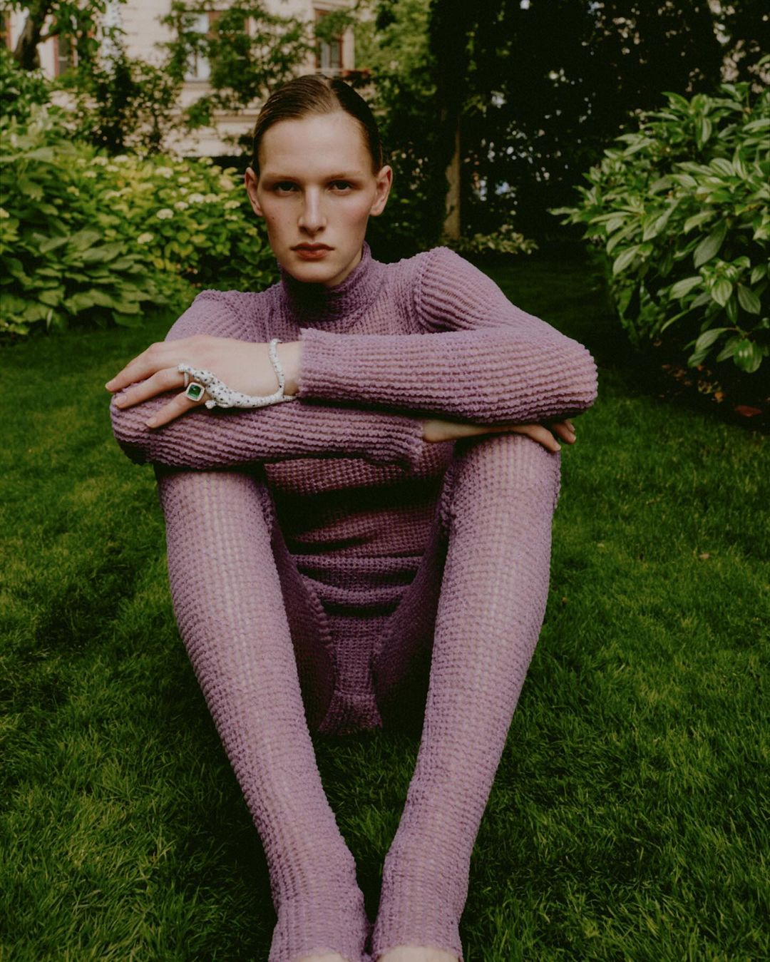 A person in a violet turtleneck and leggings sits with their legs crossed on a grass lawn outdoors near leafy green bushes and building.