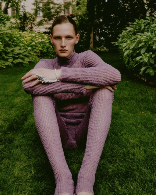 A person in a violet turtleneck and leggings sits with their legs crossed on a grass lawn outdoors near leafy green bushes and building.