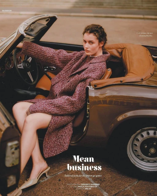 Fashion shot of a woman in a reddish purple coat sitting in the driver's seat of a brown convertible car, wearing tan mules.