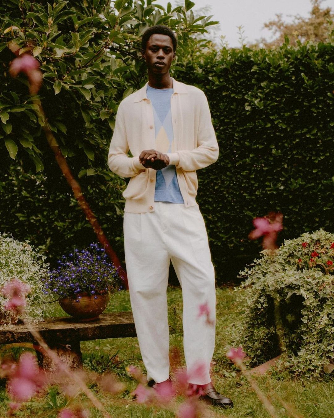 Black man standing in a lush garden wearing a cream cardigan, light blue patterned knit top, and white trousers.