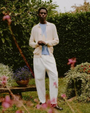 Black man standing in a lush garden wearing a cream cardigan, light blue patterned knit top, and white trousers.