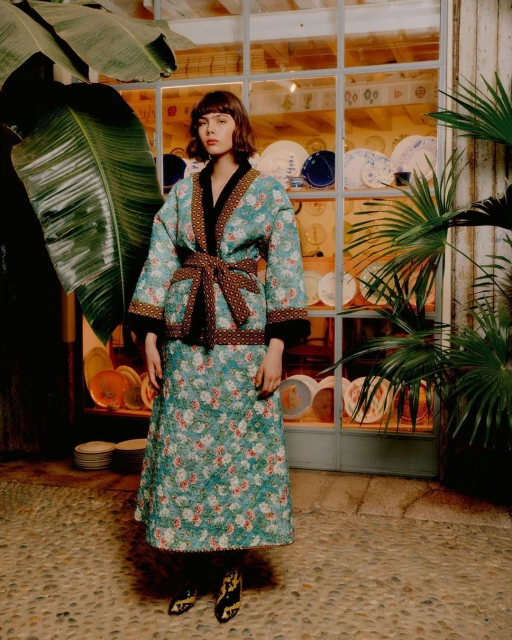 Adult female wearing a floral quilted jacket and skirt with printed boots in front of a shopfront and plants, in daylight.