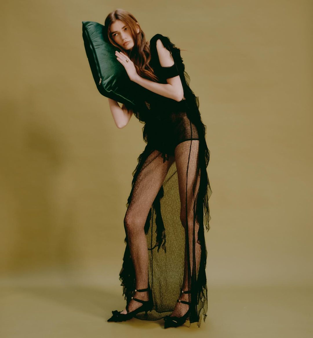 Woman in sheer black dress poses with green pillow against neutral backdrop in fashion photography. Full-length portrait, studio lighting.