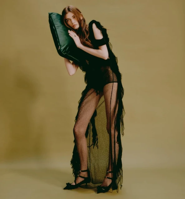 Woman in sheer black dress poses with green pillow against neutral backdrop in fashion photography. Full-length portrait, studio lighting.