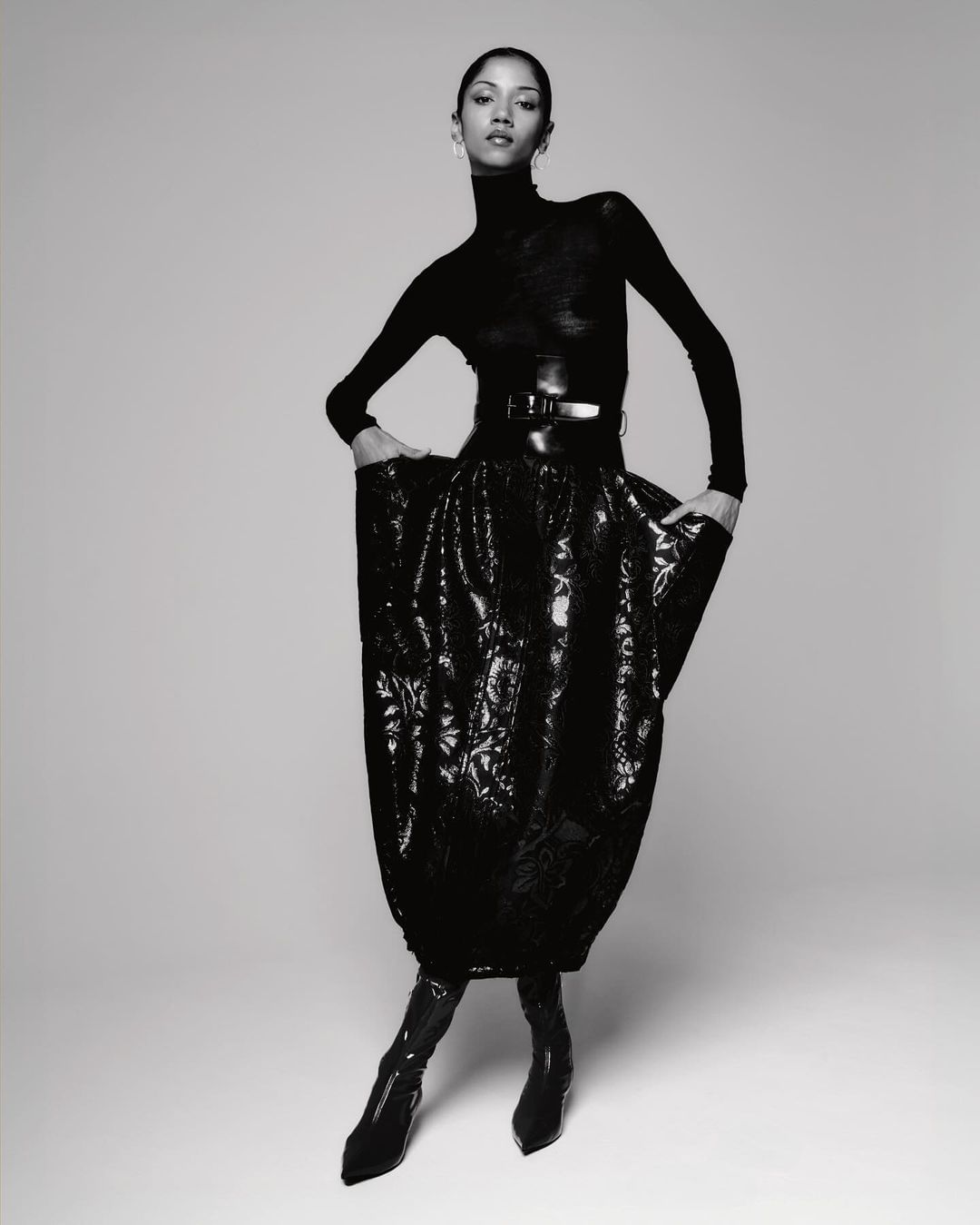 Monochrome fashion shot of a slender woman in a black turtleneck, belt, and floral skirt, standing in a photo studio.