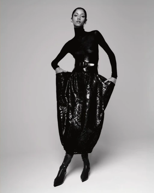 Monochrome fashion shot of a slender woman in a black turtleneck, belt, and floral skirt, standing in a photo studio.