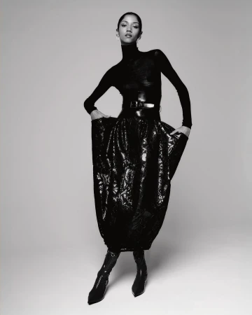 Monochrome fashion shot of a slender woman in a black turtleneck, belt, and floral skirt, standing in a photo studio.