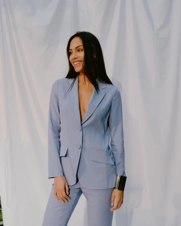 A woman with long dark hair smiles in a light blue pantsuit with a cuff bracelet, against a white draped background in a studio shot.