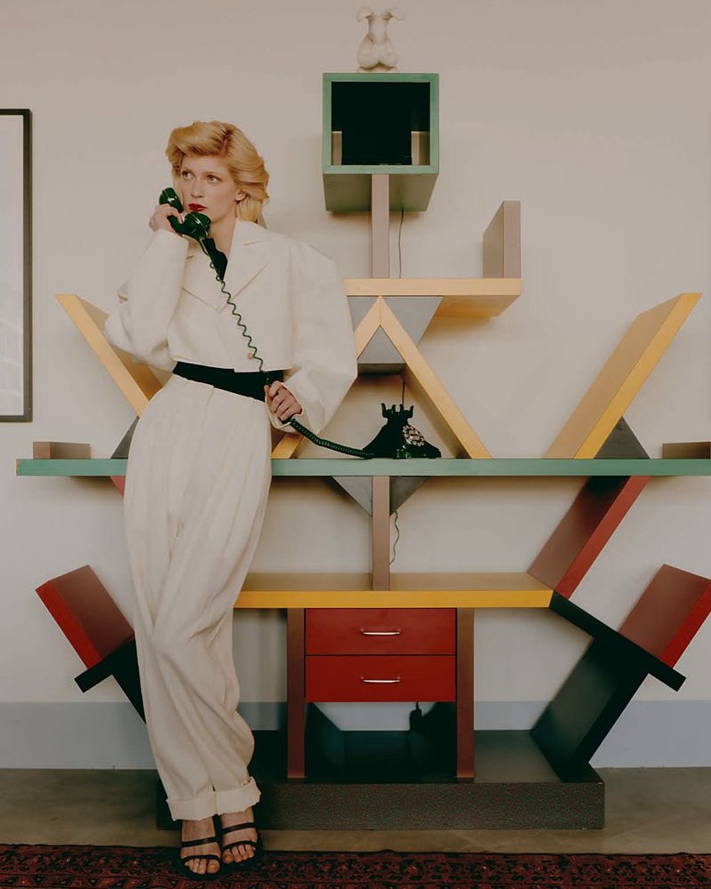 Woman in white pantsuit on phone in front of geometric bookshelf, styled in editorial fashion with muted natural light and retro feel.