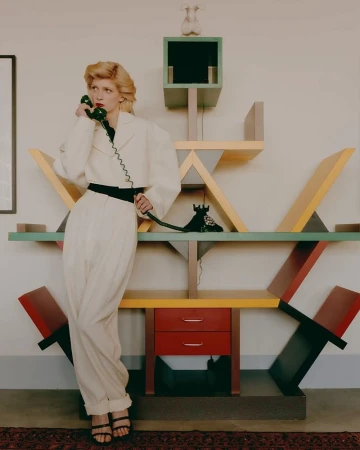 Woman in white pantsuit on phone in front of geometric bookshelf, styled in editorial fashion with muted natural light and retro feel.