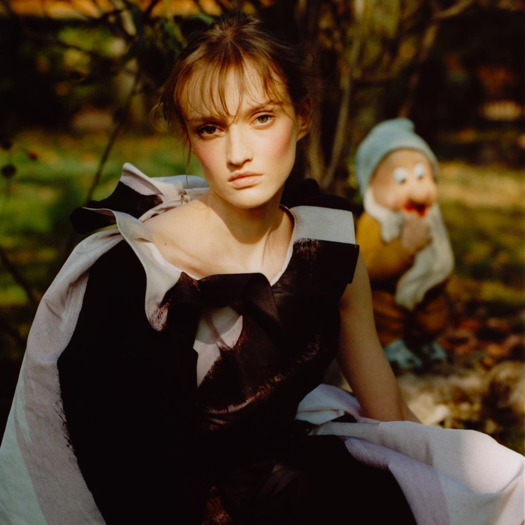 A female model with brown hair and striking makeup poses in an abstract black and white dress in a green garden, beside a gnome statue.