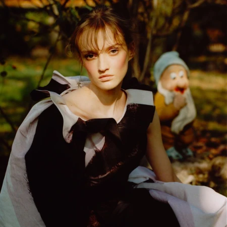 A female model with brown hair and striking makeup poses in an abstract black and white dress in a green garden, beside a gnome statue.