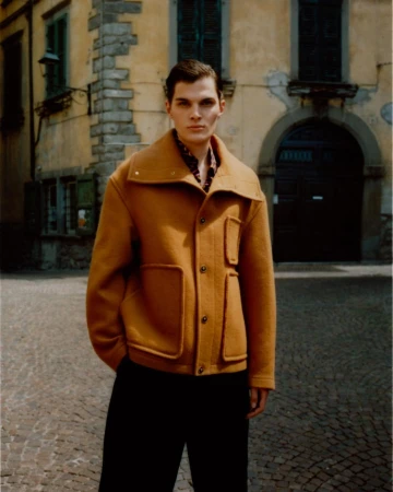 A man wearing a mustard yellow jacket and dark trousers standing on a cobblestone street in front of an aged yellow building.