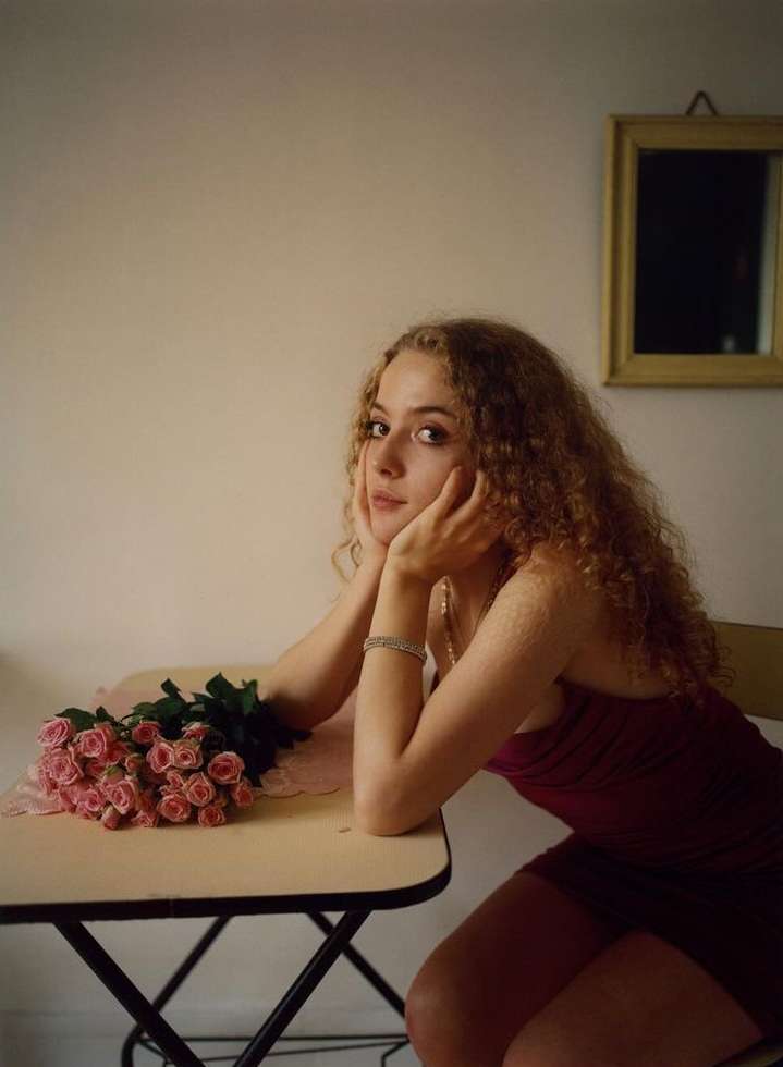Young woman with curly hair sits at a table with a bouquet of pink roses, framed by a simple interior and soft, natural lighting.