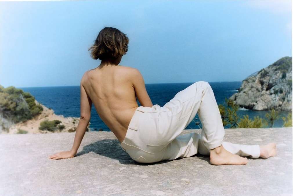 A person sits on a concrete surface overlooking the ocean. They are wearing light-colored pants and their back is to the camera.