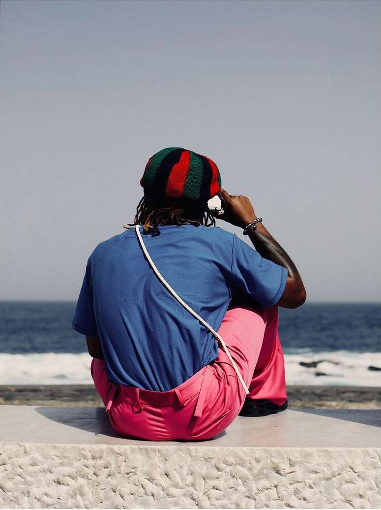 Man with dreadlocks sits facing the ocean wearing a striped hat, blue t-shirt, pink pants, and earphones. He is sitting on a stone bench.