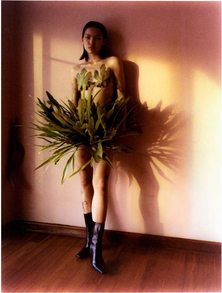 Asian woman poses with leaves and boots in a studio setting, lit by sunlight casting shadows on a pink wall, creating a natural and stylized look.