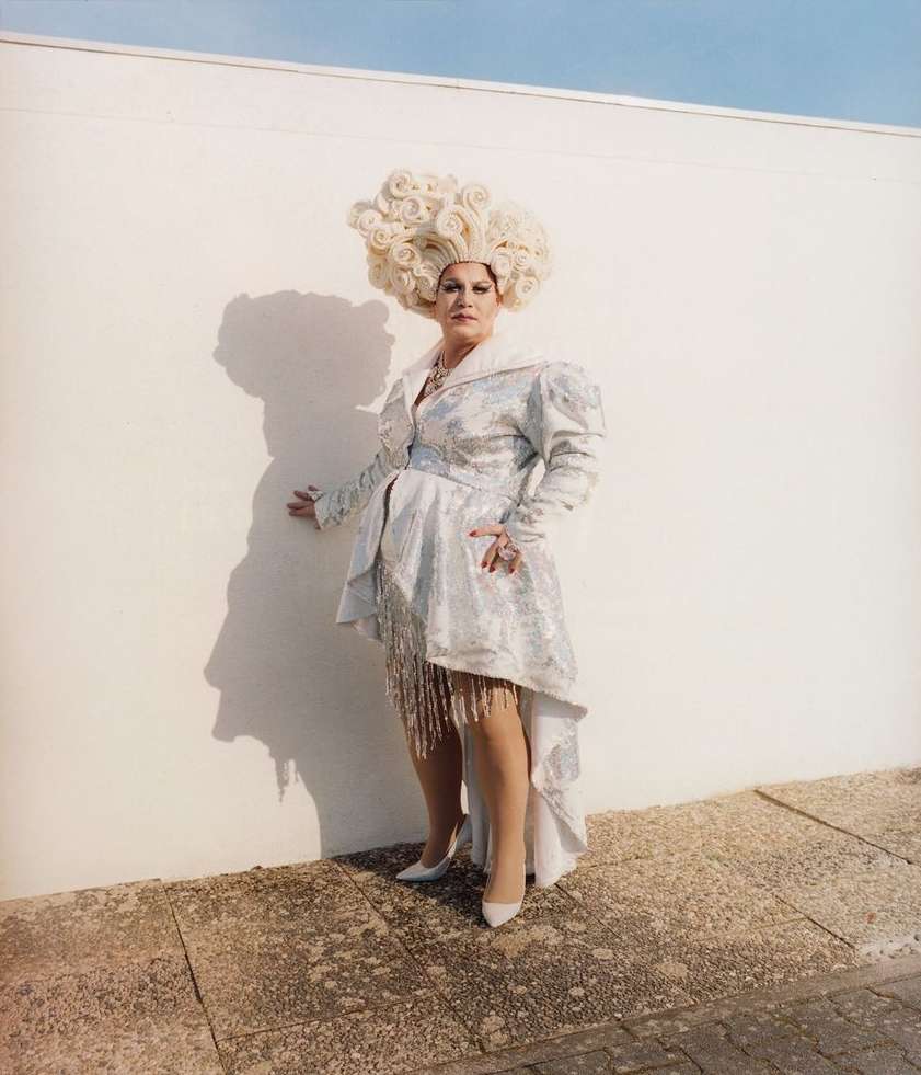 Adult female stands outdoors in ornate, high-fashion dress and exaggerated wig, posing against white wall with a neutral facial expression.