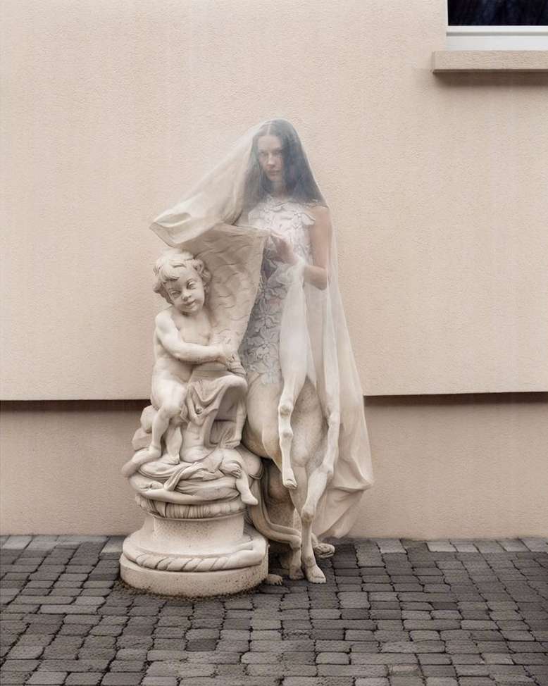 A slender woman in a white dress stands beside a sculpture of a child, partially veiled by a white cloth in a serene outdoor setting.