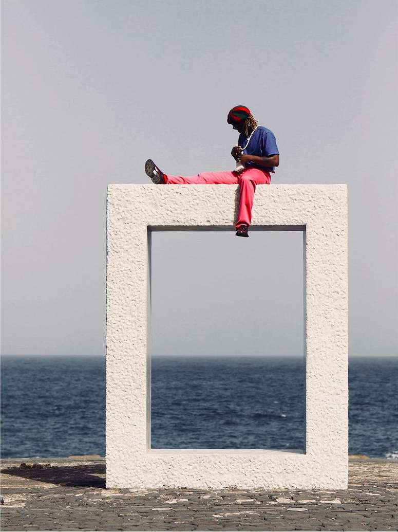 Adult male sitting atop a white concrete frame against a seascape backdrop. He wears a hat, blue shirt, and pink pants in a sunny scene.