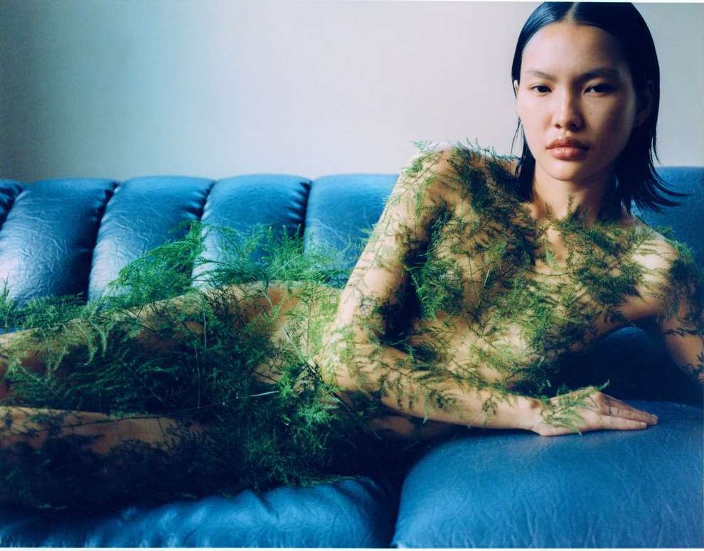 Asian woman reclines on a blue couch covered in green ferns, looking at the camera in an indoor setting.