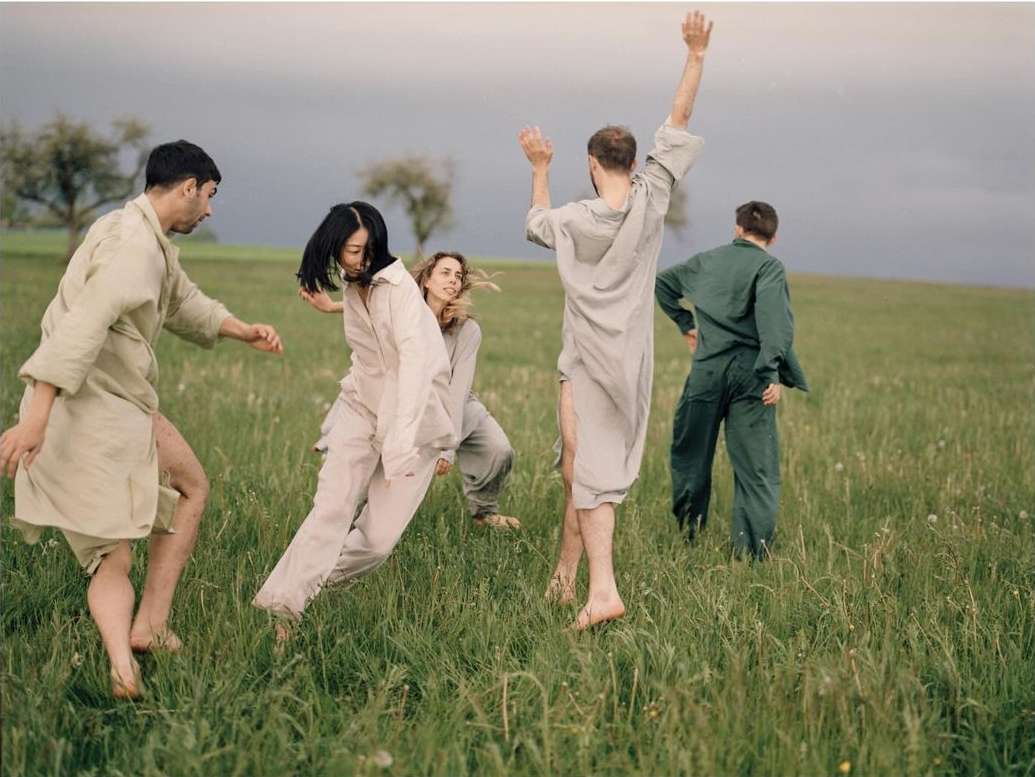 Five barefoot people in loose clothing gather in a grassy field under a cloudy sky, with trees in the background, evoking a natural, carefree mood.
