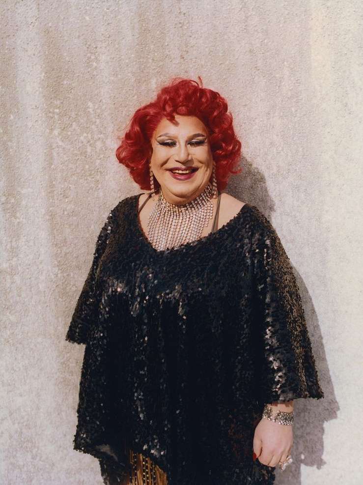 Portrait of an adult woman with bright red hair and a sparkly black poncho-style top standing against a neutral wall indoors.