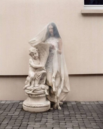 Woman wearing a sheer veil standing next to a classical stone cherub statue against a textured beige exterior wall