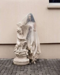 Woman wearing a sheer veil standing next to a classical stone cherub statue against a textured beige exterior wall