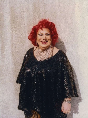 Portrait of a smiling older individual with vibrant red hair wearing a sparkly black top and heavy crystal necklace against a textured wall.