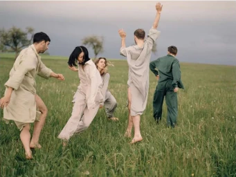 Group of five people in loose clothing running and dancing barefoot across a green grassy meadow under an overcast sky.