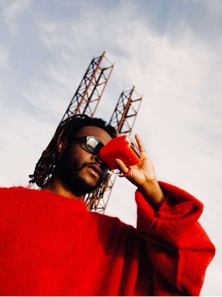 Low-angle shot of a man wearing a bright red sweater and sunglasses, holding a small red device against his face outdoors.