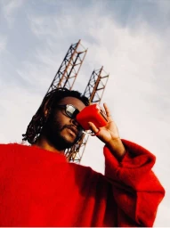 Low-angle shot of a man wearing a bright red sweater and sunglasses, holding a small red device against his face outdoors.