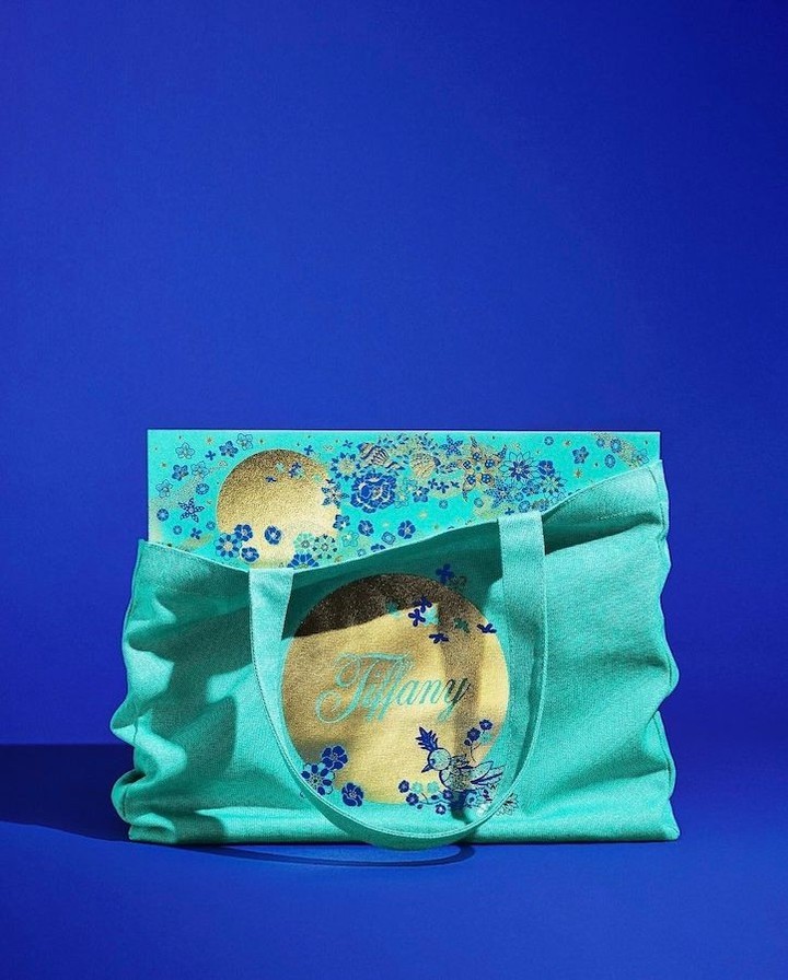 Turquoise tote bag with gold floral design and a Tiffany box inside, presented against a royal blue background in a studio shot.