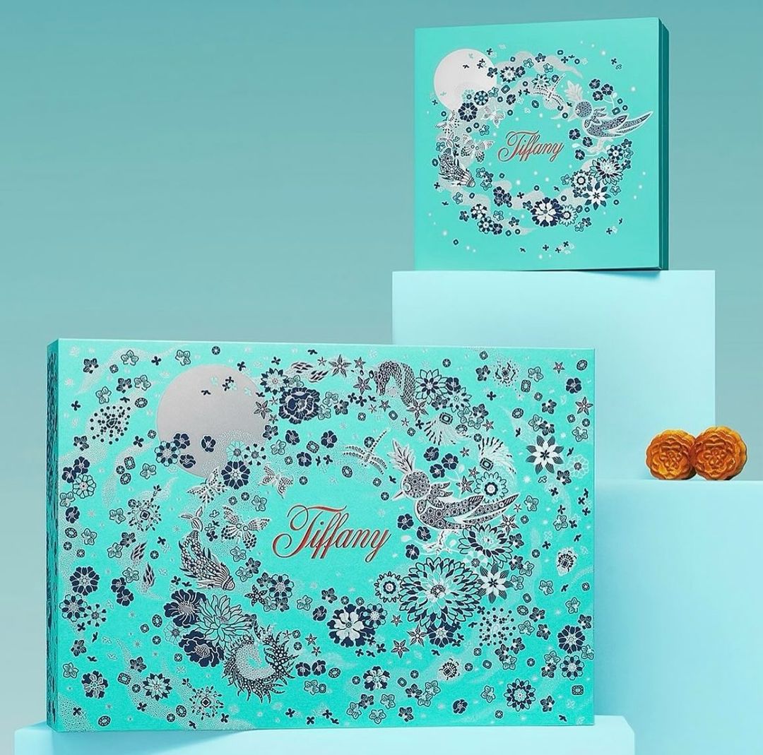 Two Tiffany & Co. branded boxes with decorative floral, moon, and bird designs on a light blue surface, with two pastry treats.
