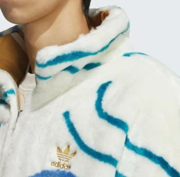 Close-up of a person wearing a white and blue faux fur jacket with an embroidered gold trefoil logo on a plain white background.
