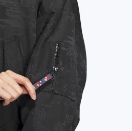 Close-up of a person pulling the zipper tab on the sleeve pocket of a black patterned bomber jacket against a plain background