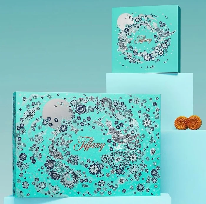Turquoise Tiffany boxes featuring intricate silver floral illustrations and typography, arranged on tiered platforms with two mooncakes.