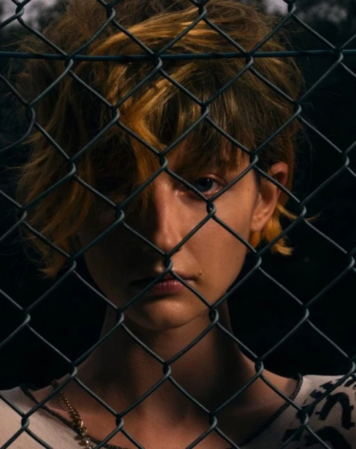 Close-up of a person with blonde-streaked hair looking through a chain-link fence against a dark background, creating a dramatic, obscured portrait.