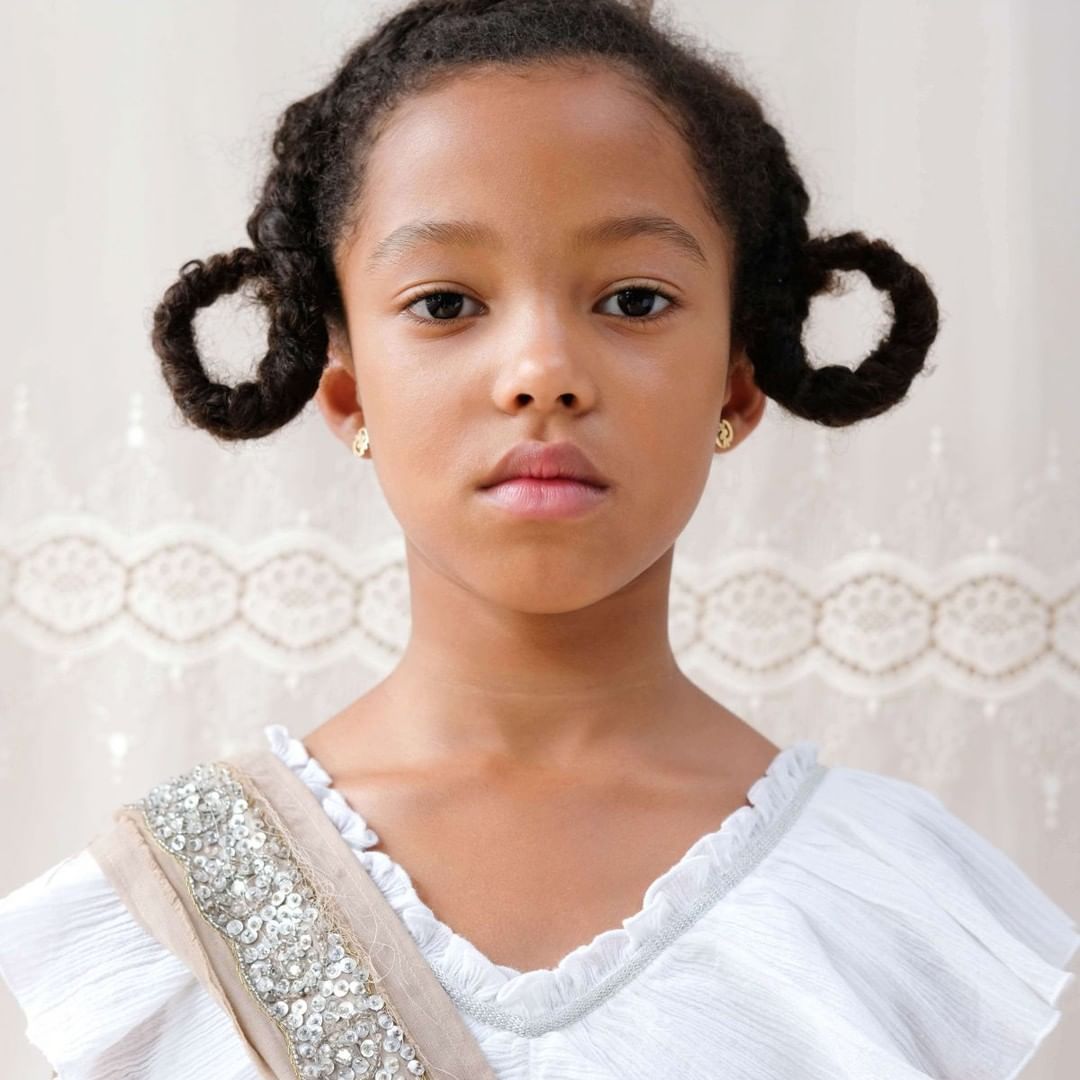 Close-up studio portrait of a girl with an earnest expression wearing a white top with a sequined sash against a neutral backdrop.