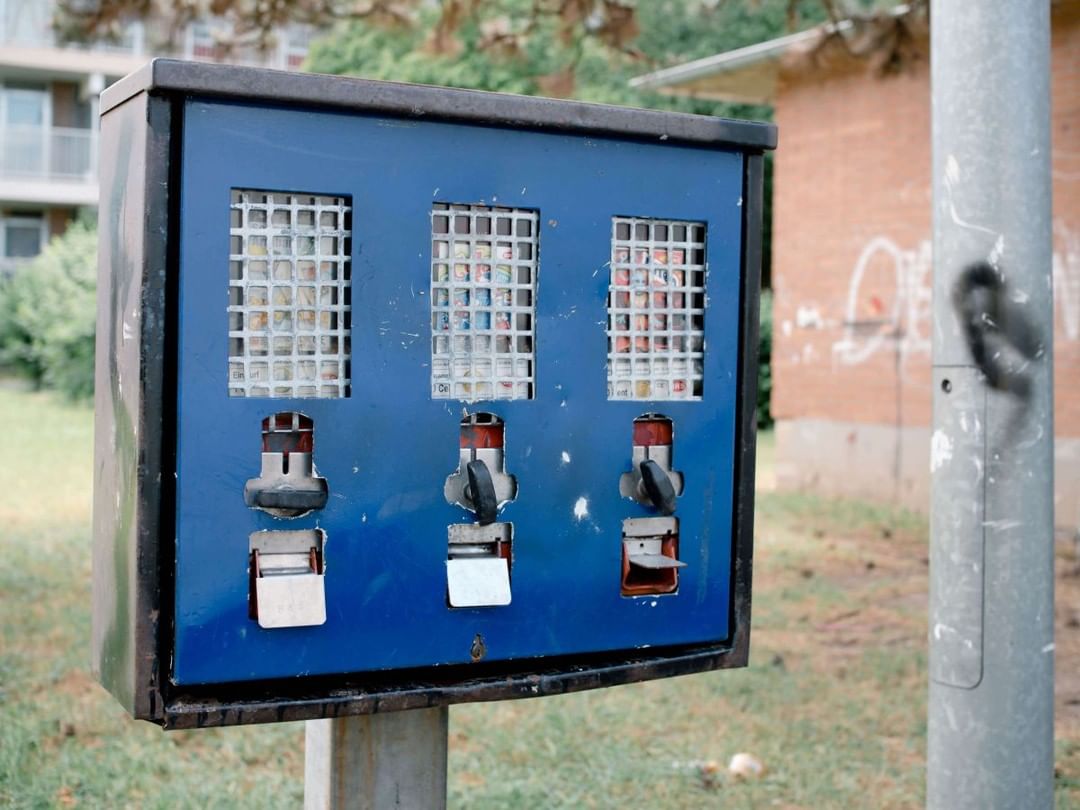 Vending machine with three dispensers outdoors. Blue metal casing with coin slots, product windows, and turn knobs. Urban background.