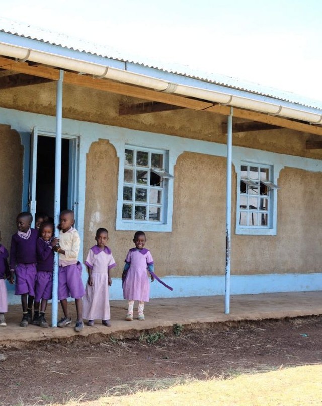 Group of young children standing outside of a school building with a blue trim. The kids are wearing various purple clothing in daylight.