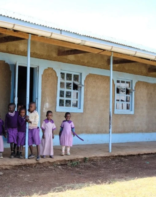 Group of young children standing outside of a school building with a blue trim. The kids are wearing various purple clothing in daylight.